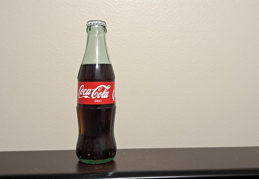 A single glass bottle of Coca-Cola, positioned on the left side of a dark, horizontal surface.